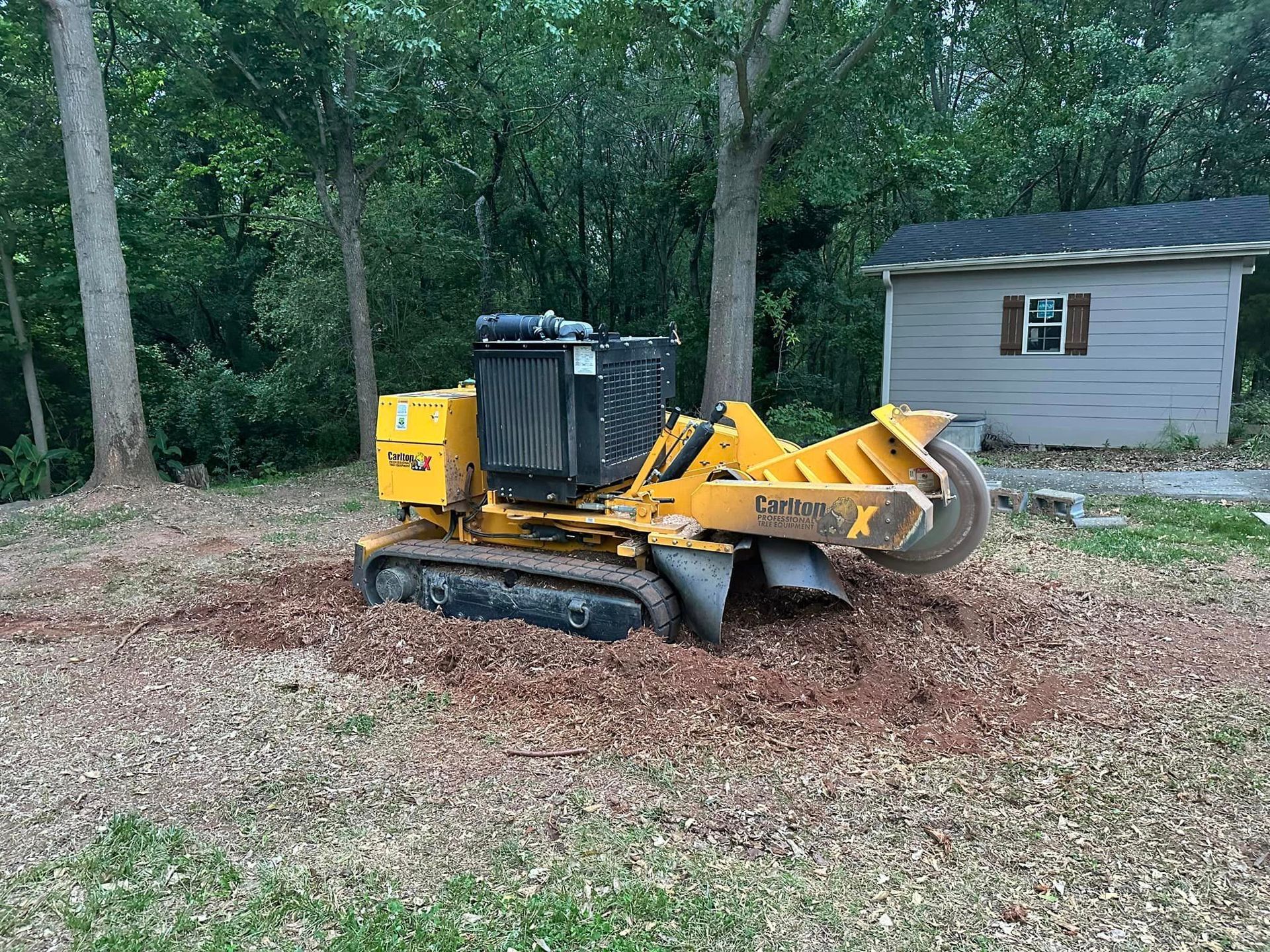 A yellow stump grinder is sitting in a field next to a house.