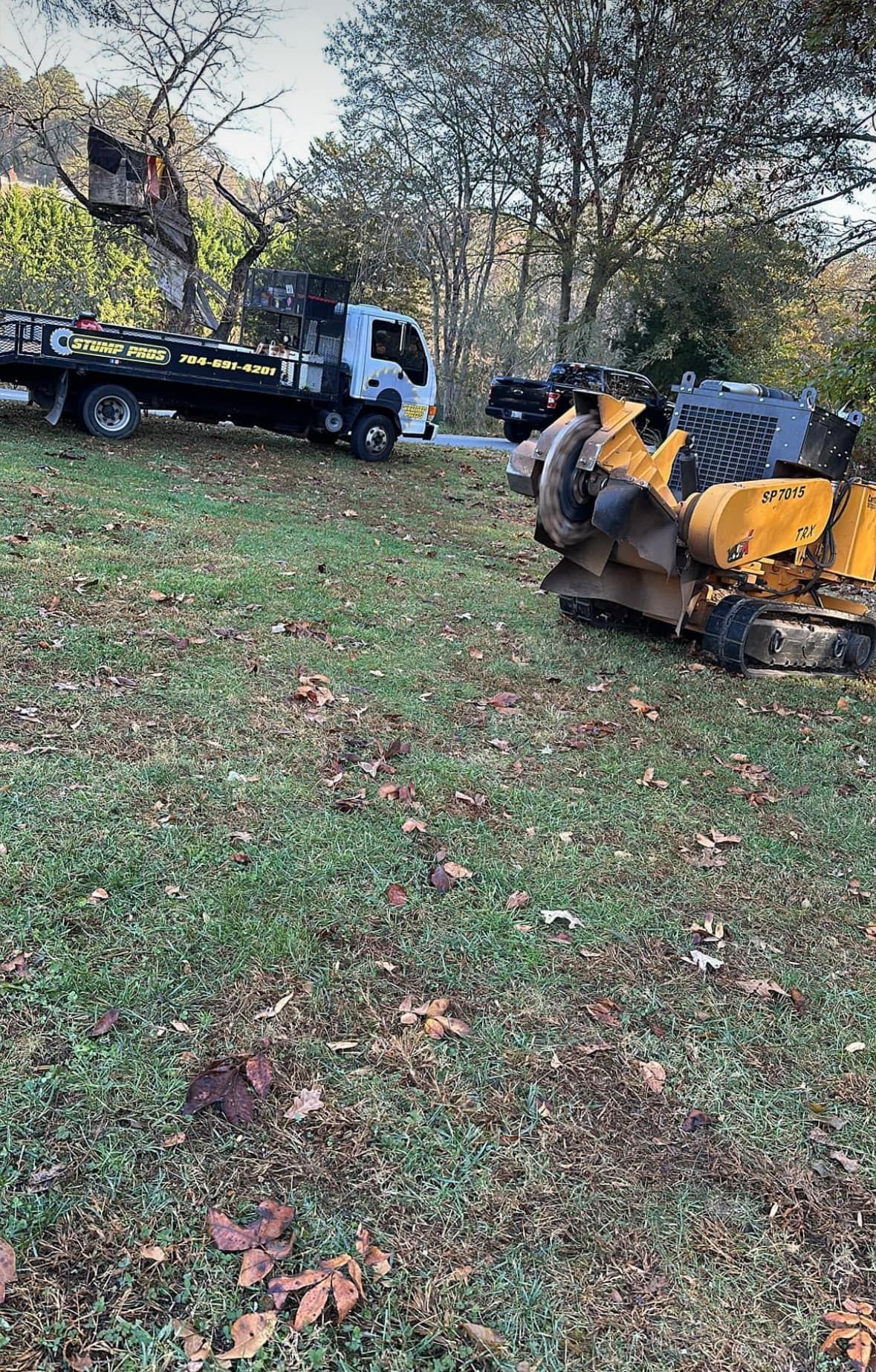 A tow truck is towing a tree stump grinder in a grassy field.