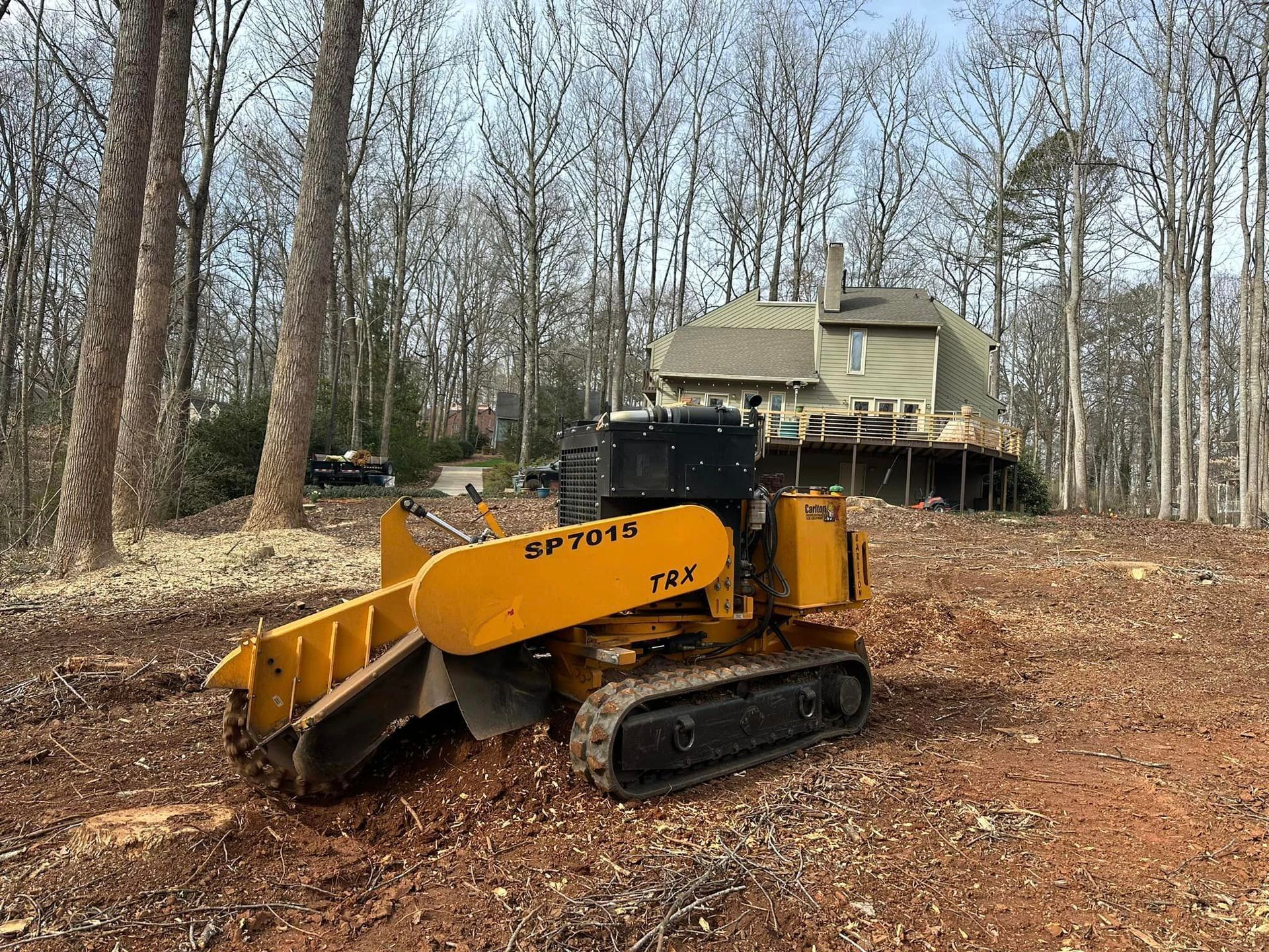 A yellow stump grinder is sitting in a dirt field in front of a house.