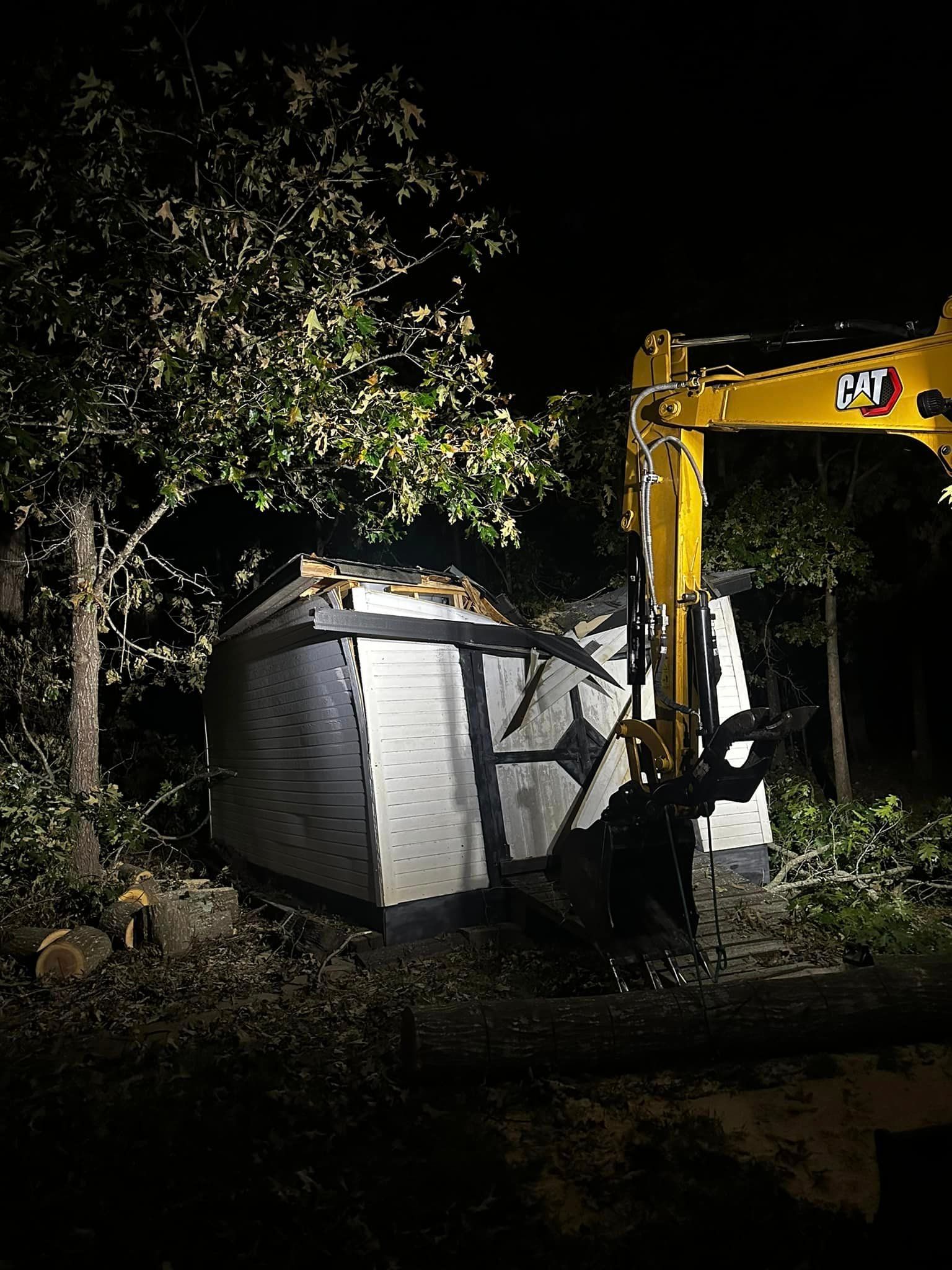 A yellow excavator is demolishing a shed at night.