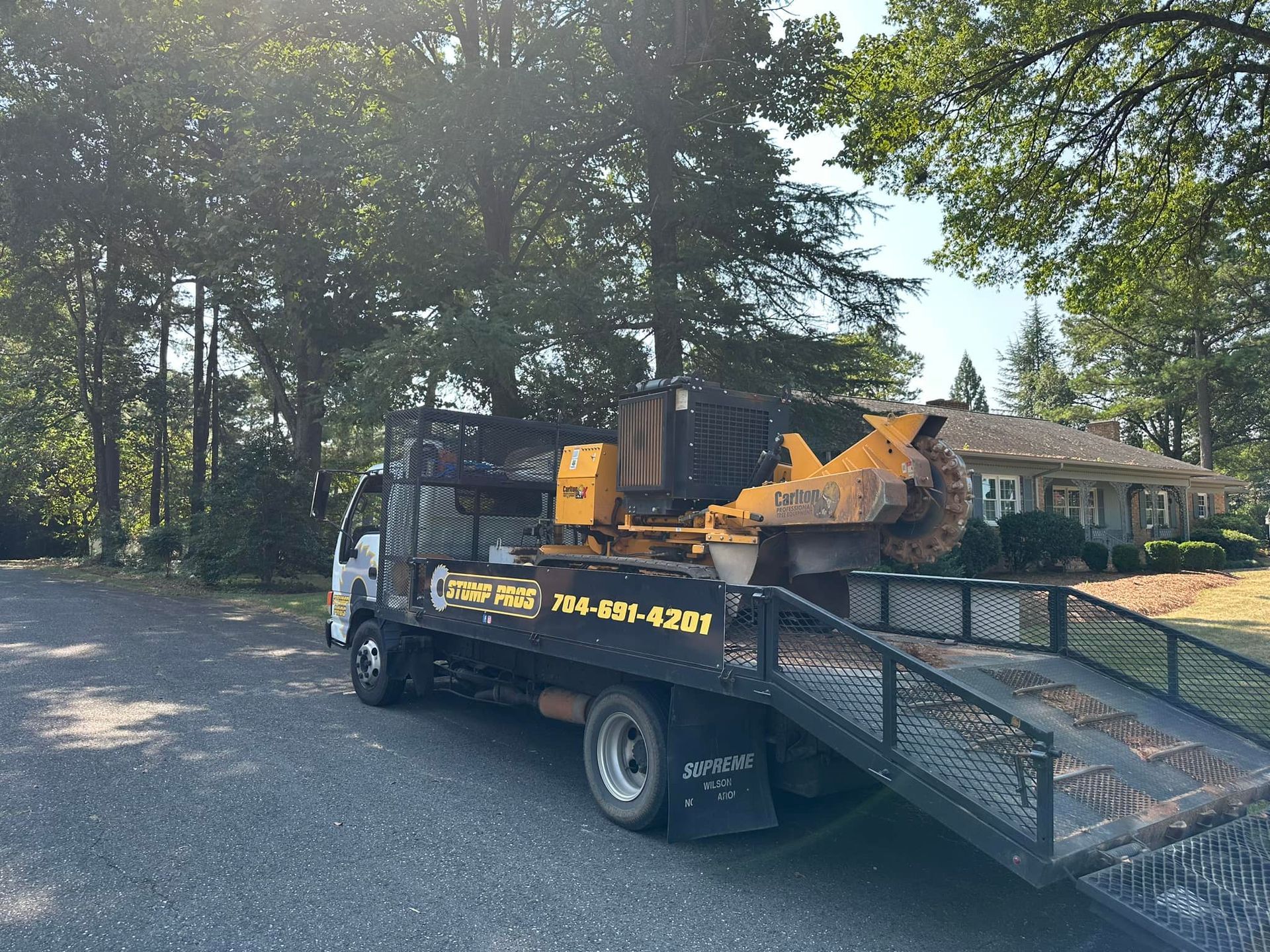 A tow truck with a stump grinder on the back is parked in front of a house.