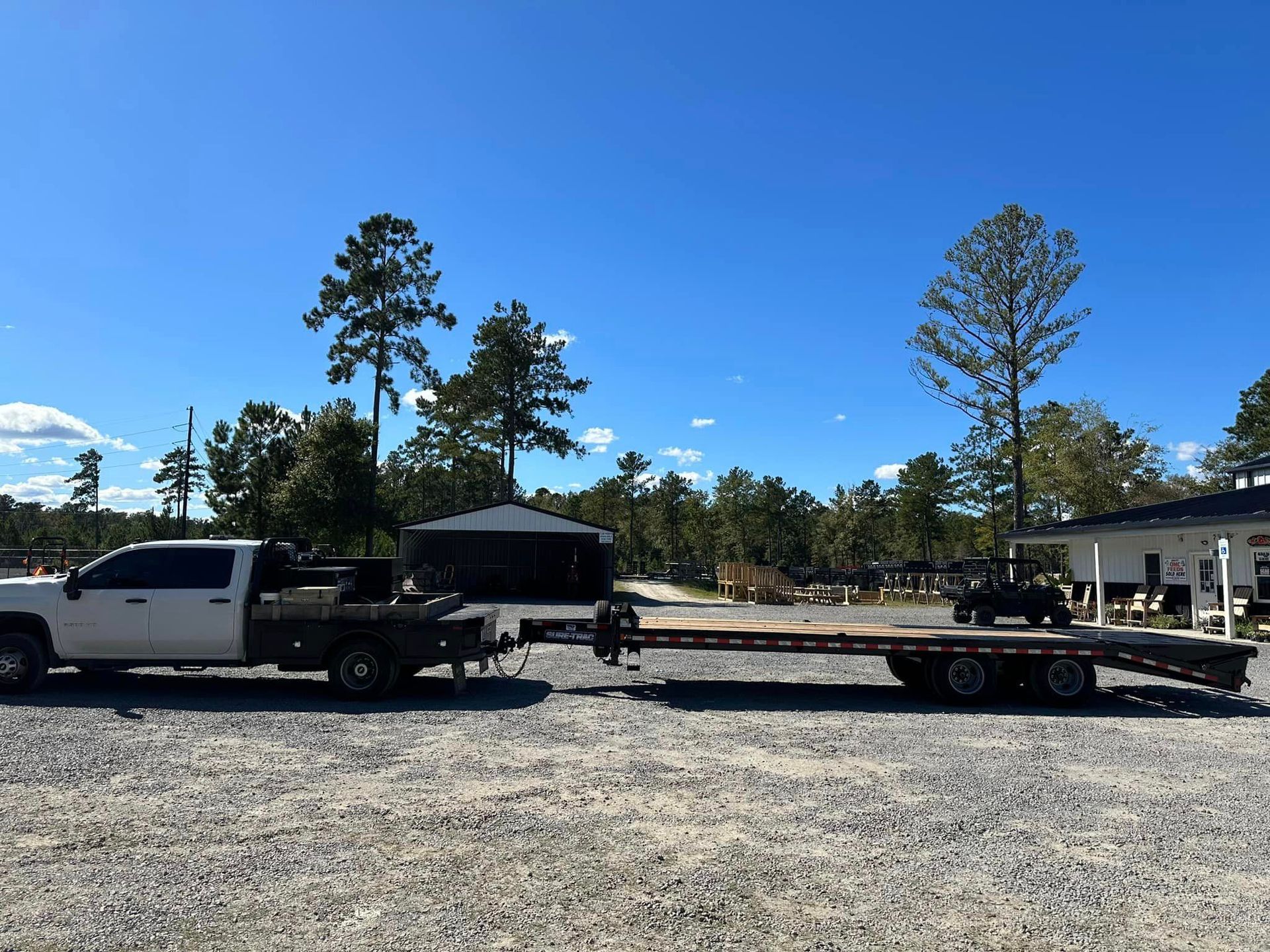A white truck is towing a flatbed trailer in a gravel lot.