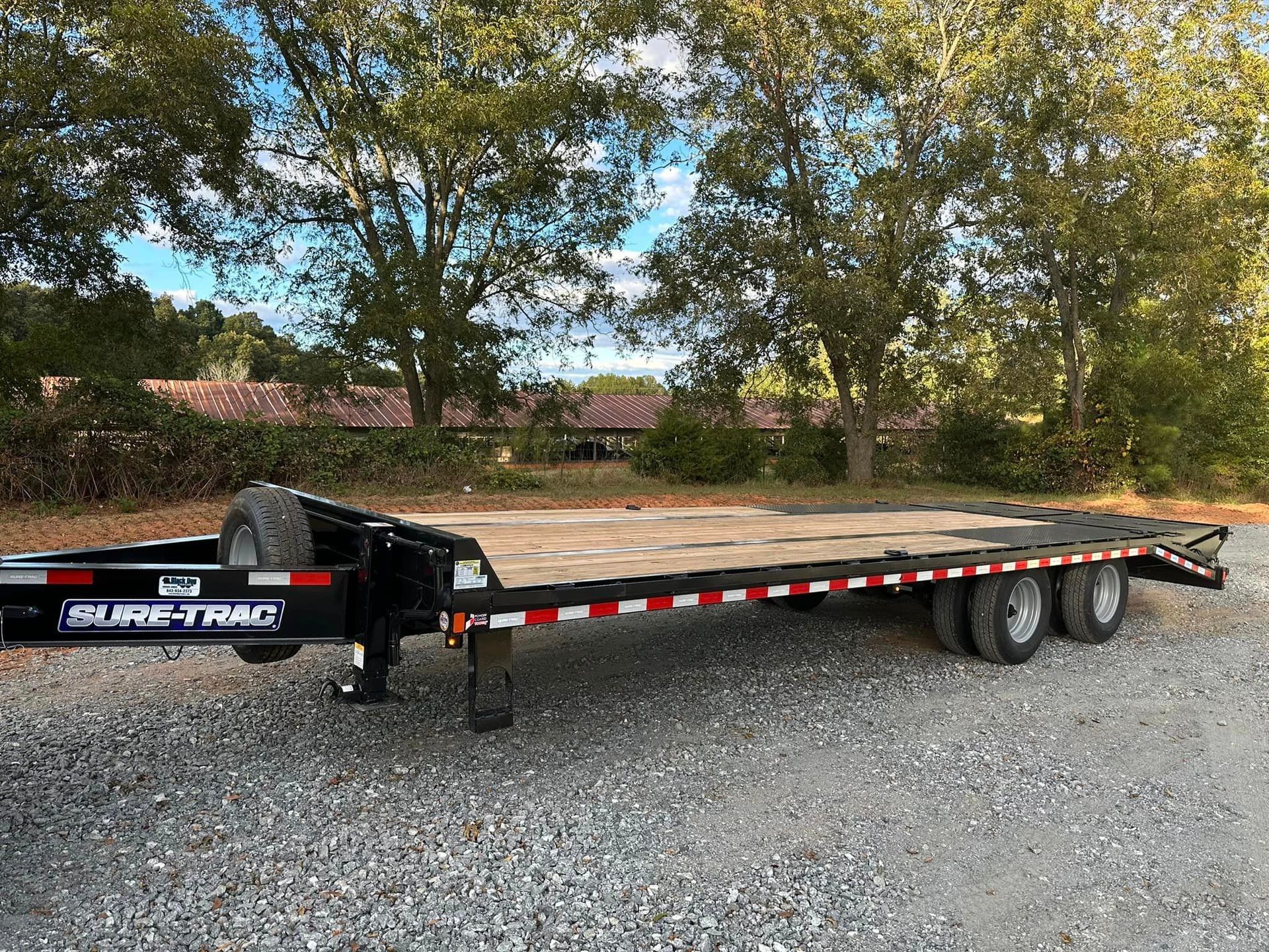 A flatbed trailer is parked in a gravel lot with trees in the background.