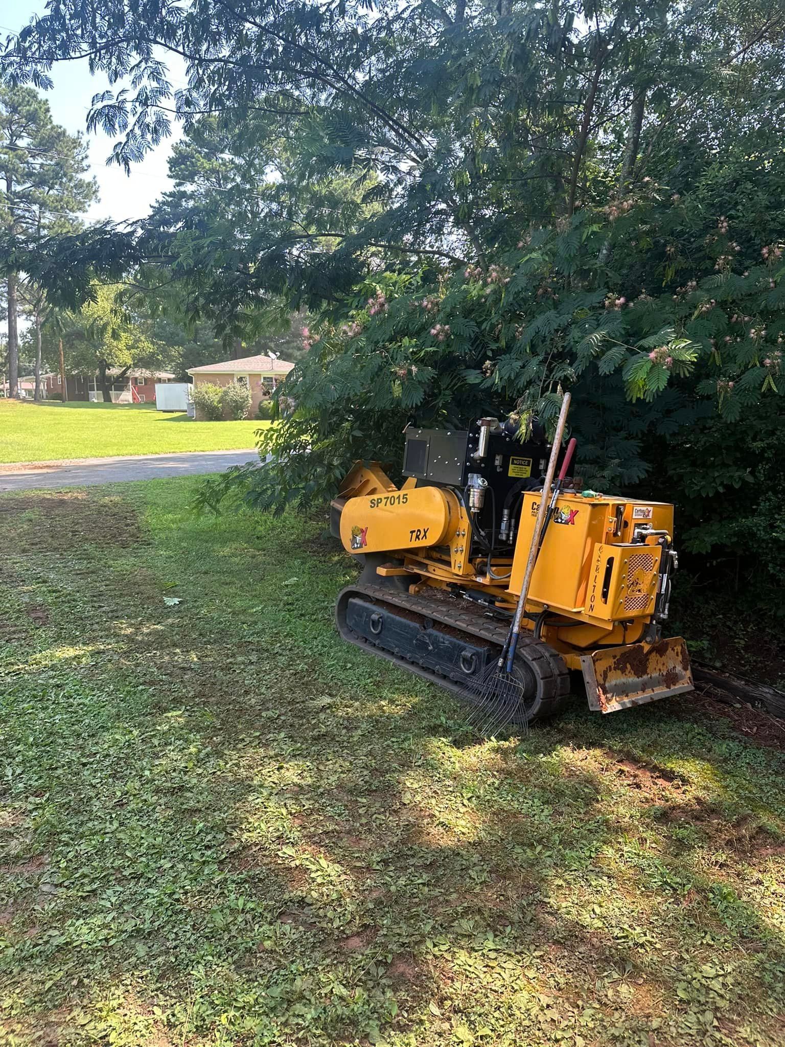 A yellow stump grinder is sitting in the grass next to a tree.