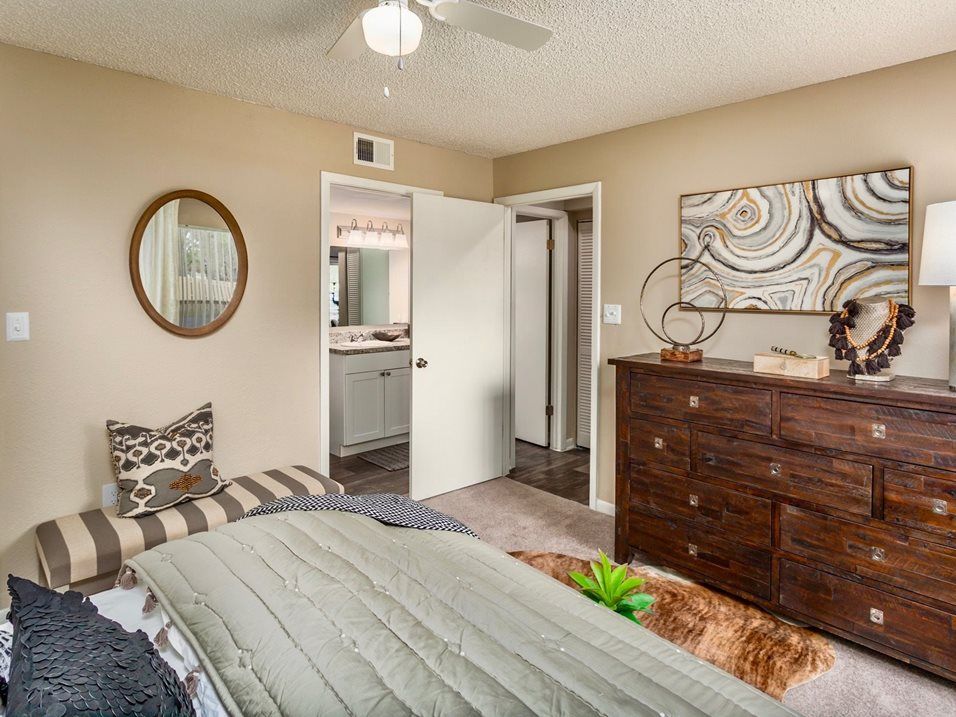 Bedroom with a bed, dresser, mirror, and a partially open door to a bathroom. Beige walls, neutral colors.