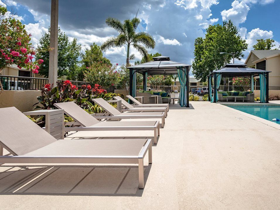 Lounge chairs by a pool with two shaded cabanas, under a cloudy sky.