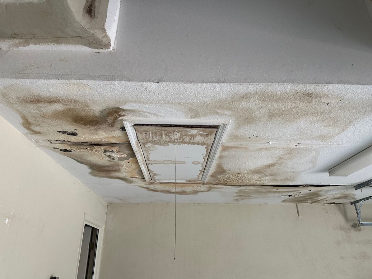 A garage ceiling with large, dark water stains and peeling texture surrounding a closed attic access panel.