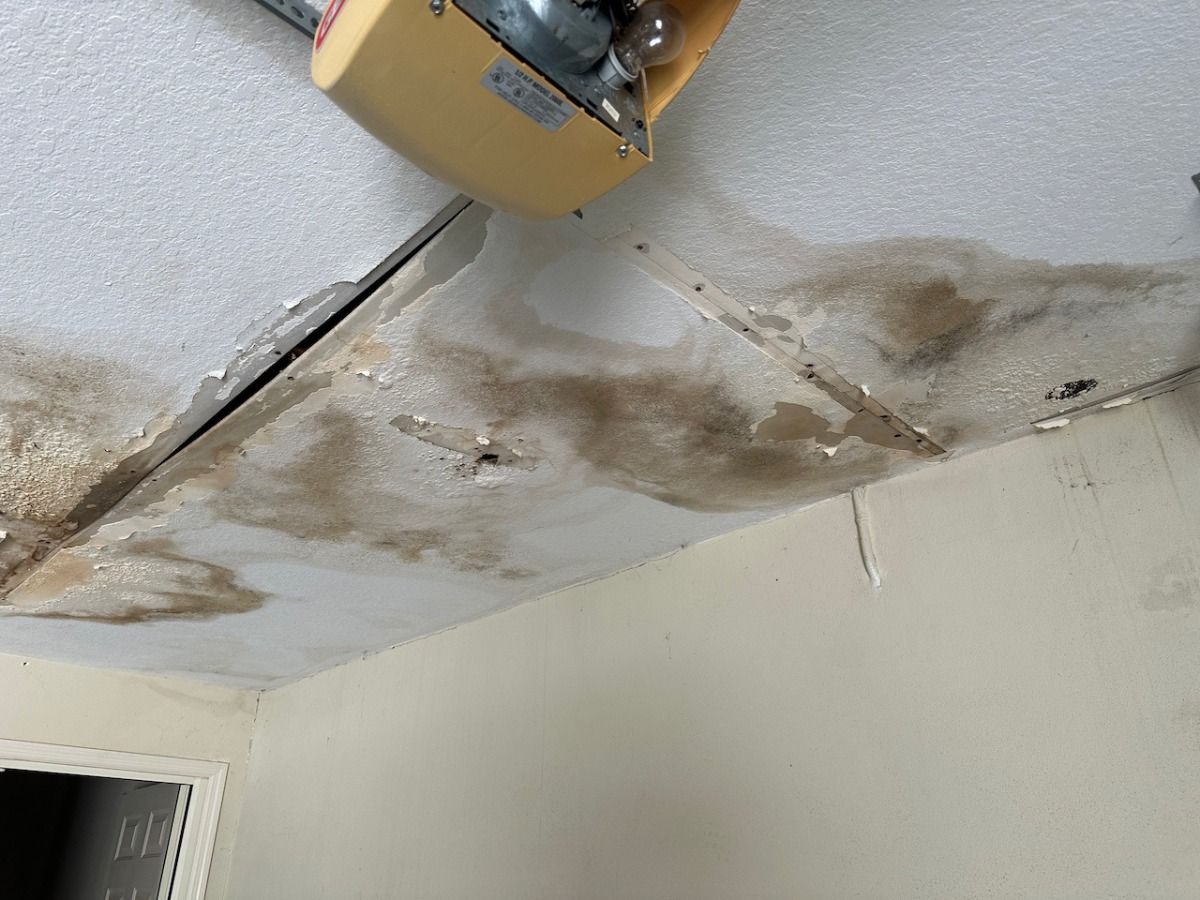 A damaged garage ceiling showing large brown water stains, peeling paint, and a garage door opener motor attached.