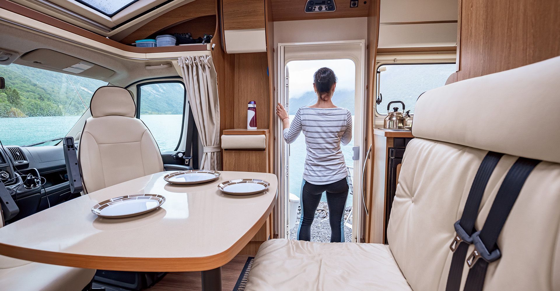 Interior of a camper, person looking out door to a body of water. Beige furniture, plates on a table.