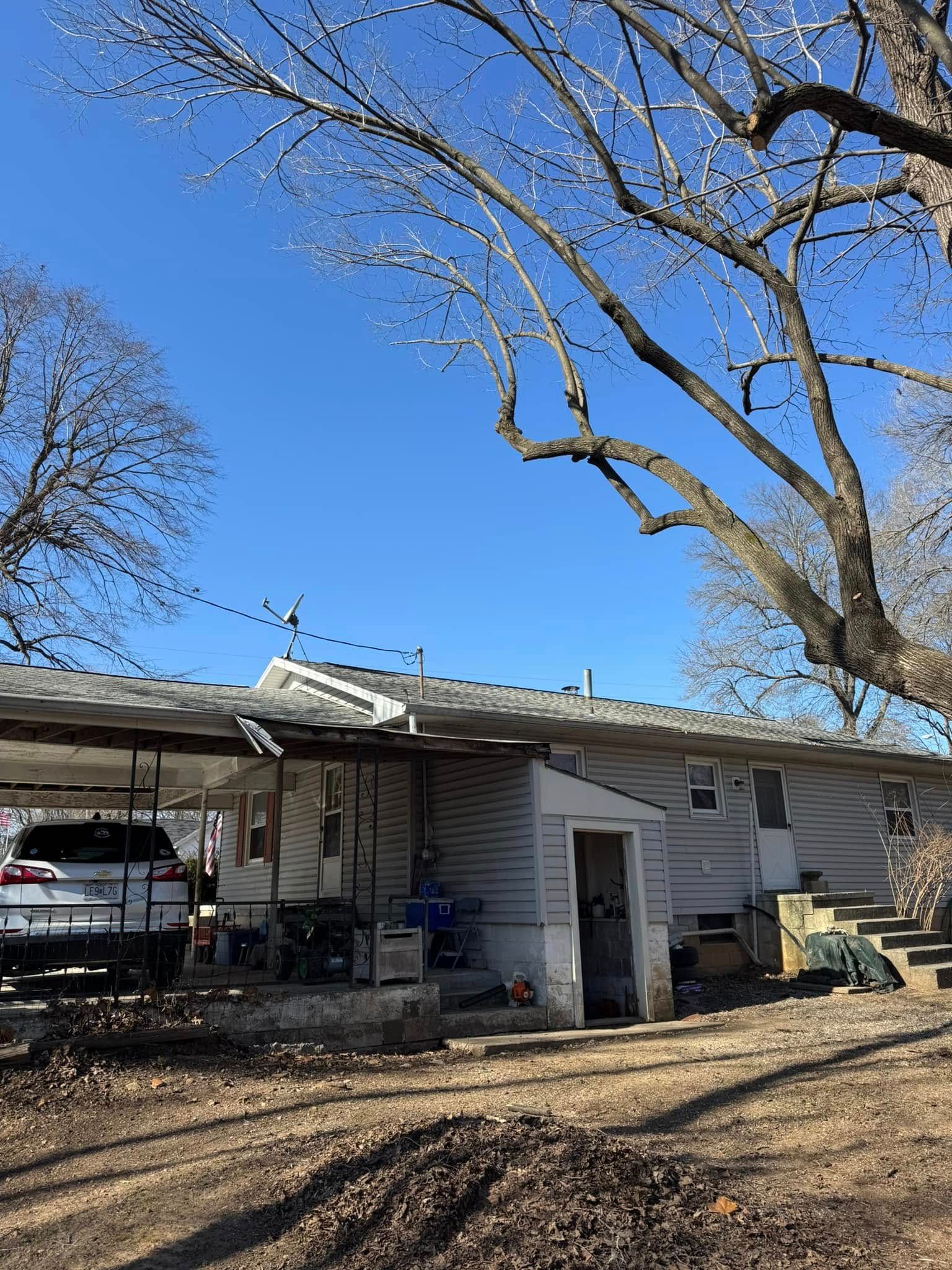 House with carport, leafless trees, bright blue sky. Car parked under carport, dirt ground.