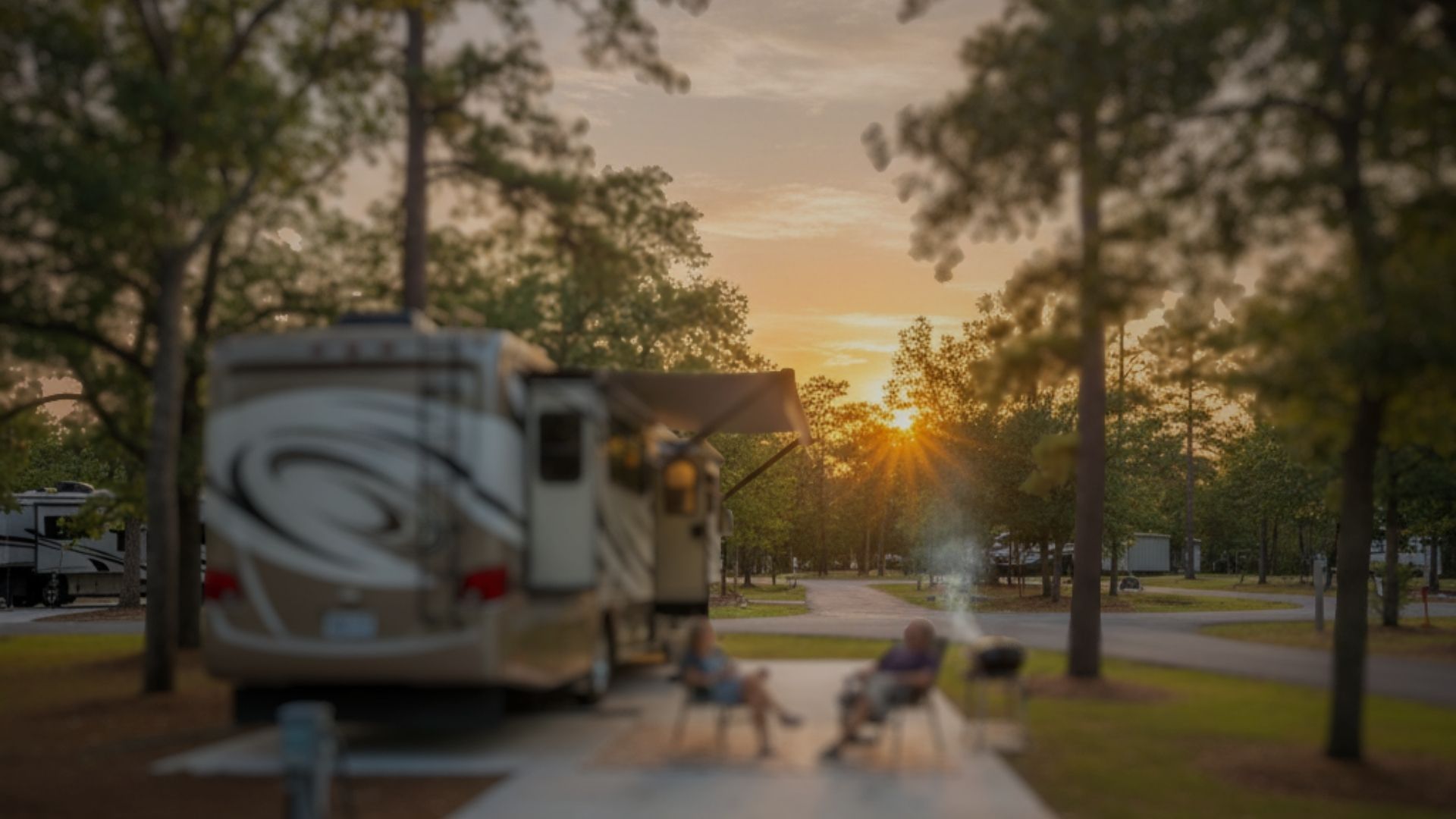 RV parked at a campsite, chairs in front, trees, sunset in background.