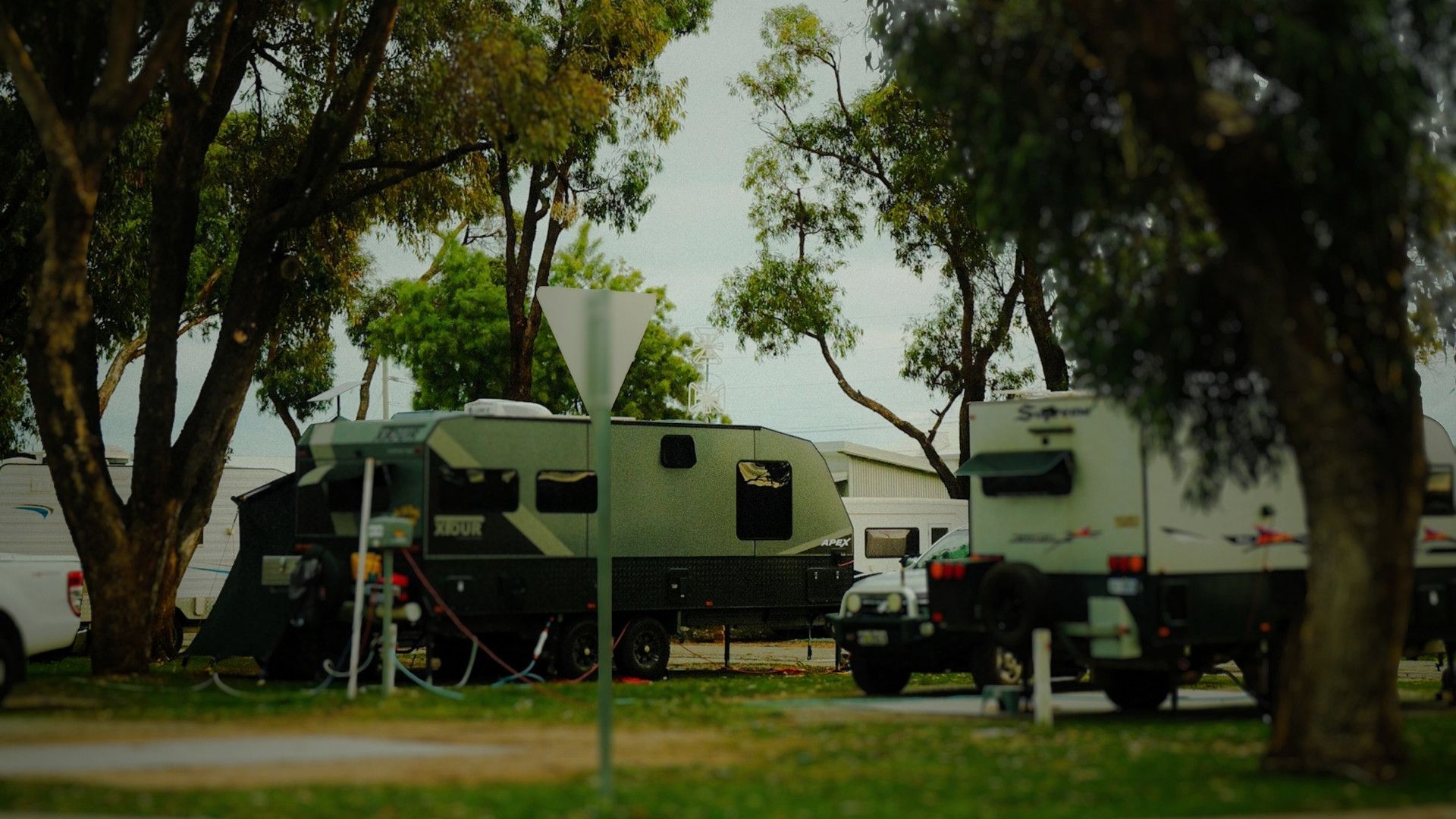 Campers and trailers under trees on a grassy area, cloudy sky in the background.