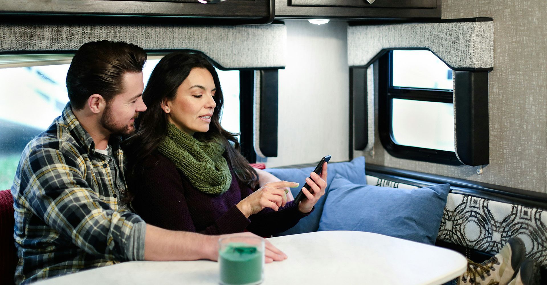 Couple in RV looking at a phone, seated at a table. Woman points at screen, man smiles.