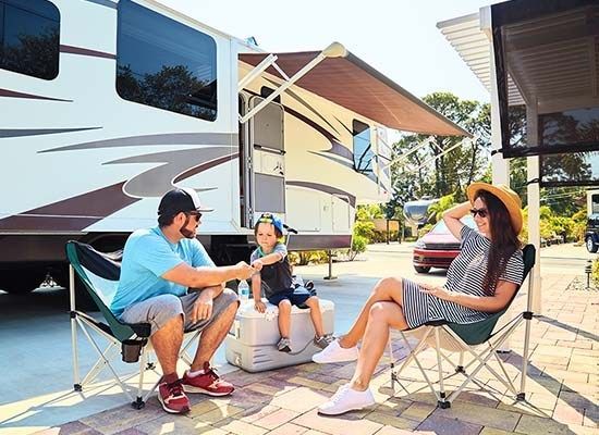 Family relaxing outside RV, under awning, in camping chairs. Sunny day.