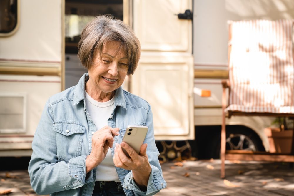 Woman in denim shirt smiles while using a phone, near a recreational vehicle outdoors.