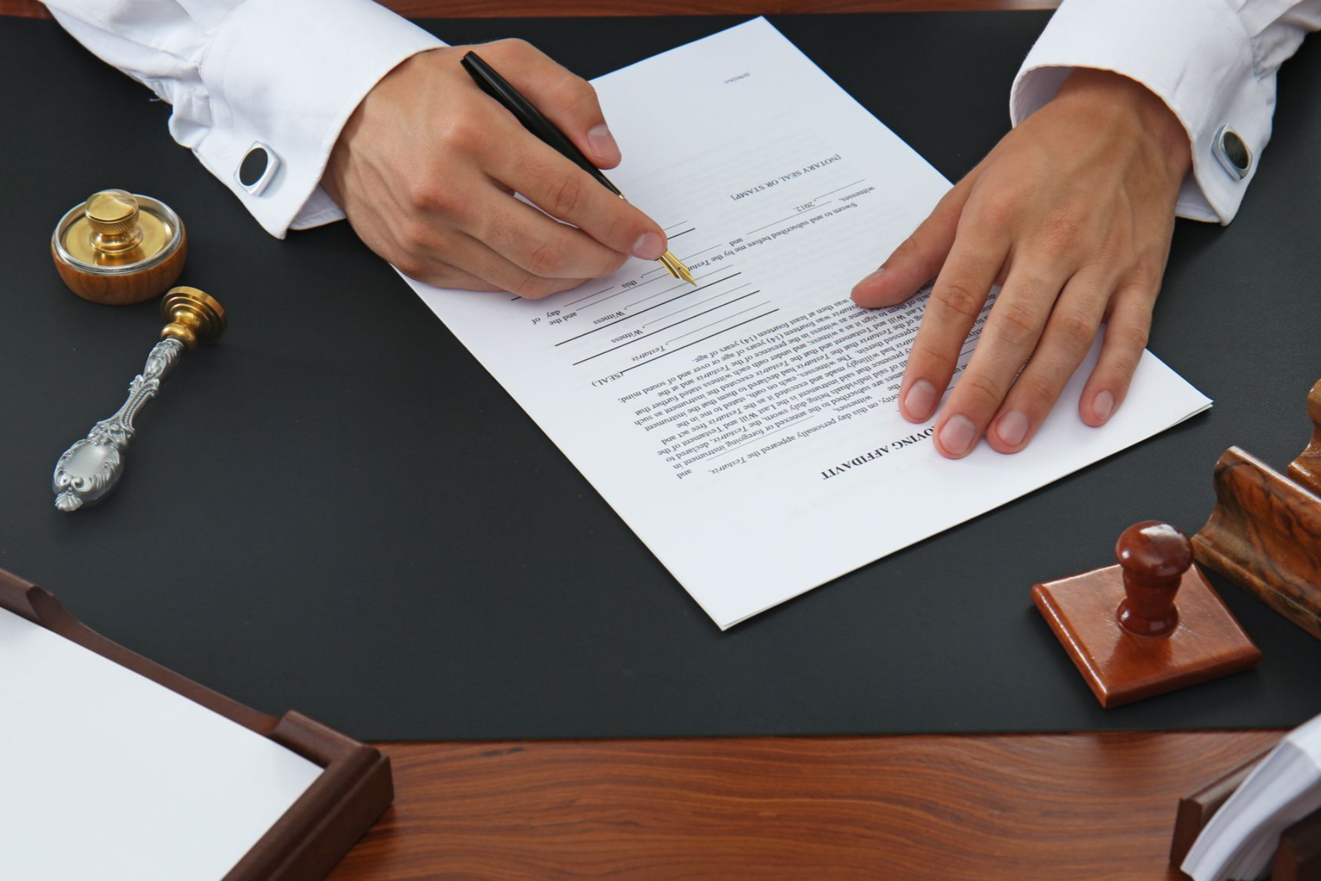 Person signing a document on a desk with stamps; formal setting, white sleeves, dark surface.