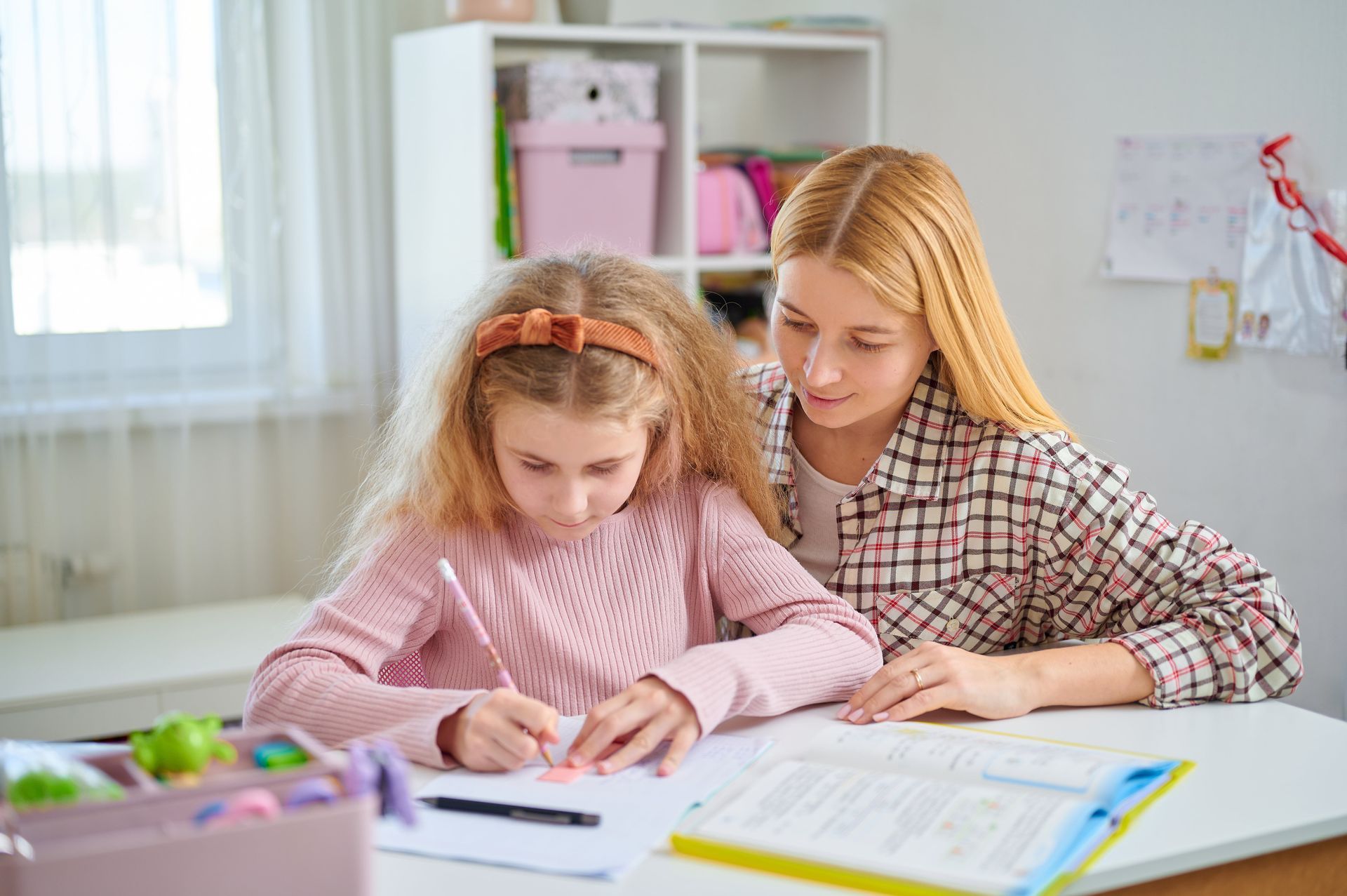 Blonde woman helping young girl write at a desk; room setting with white furniture, pink and green items.