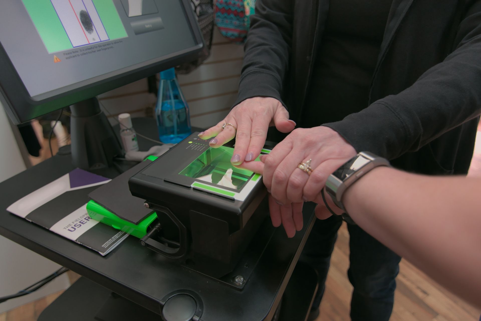 Person using a fingerprint scanner at a counter. One person's fingers are on the scanner while another person's hand assists.