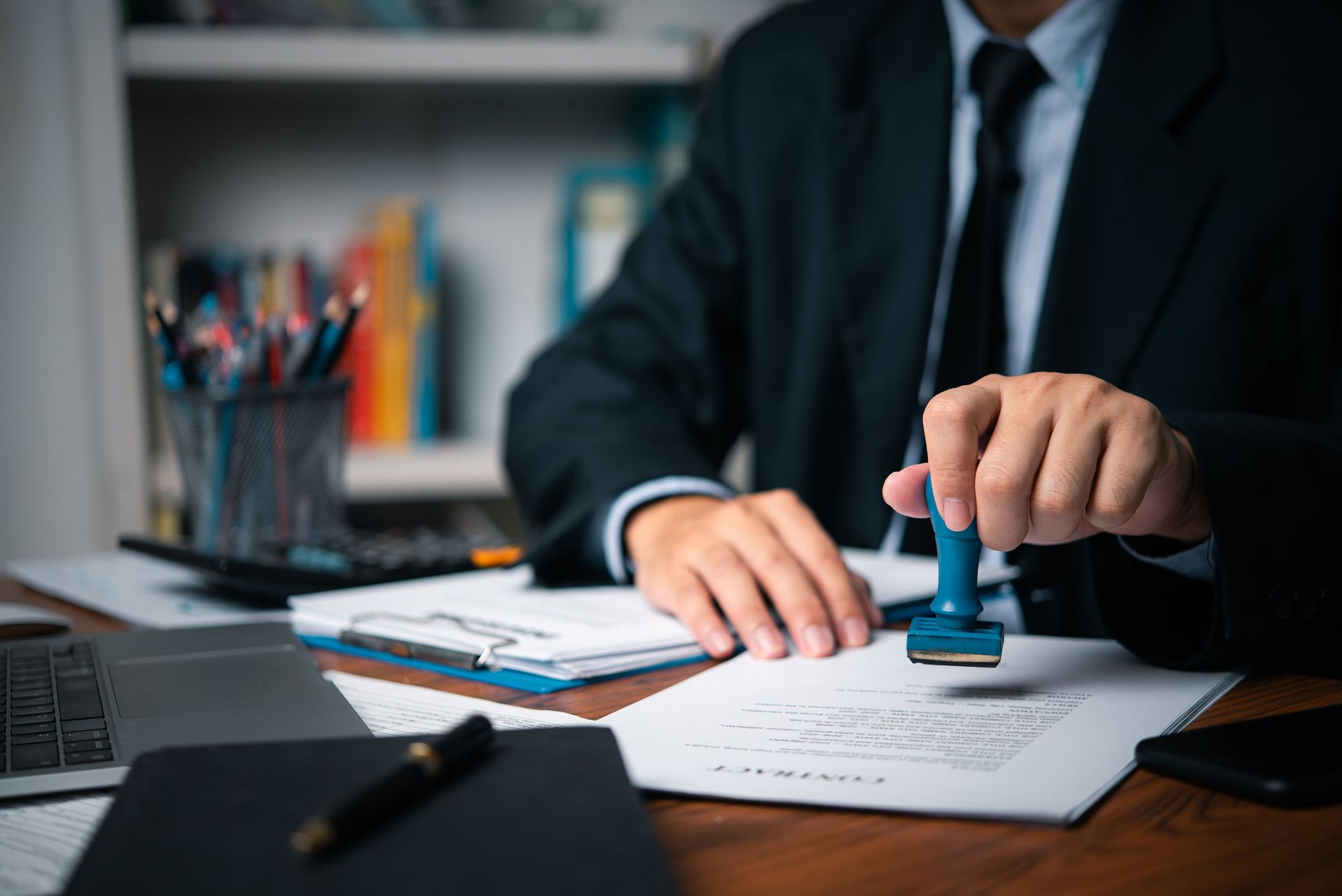 Person in a suit stamps a document with a blue stamp at a desk with office supplies.