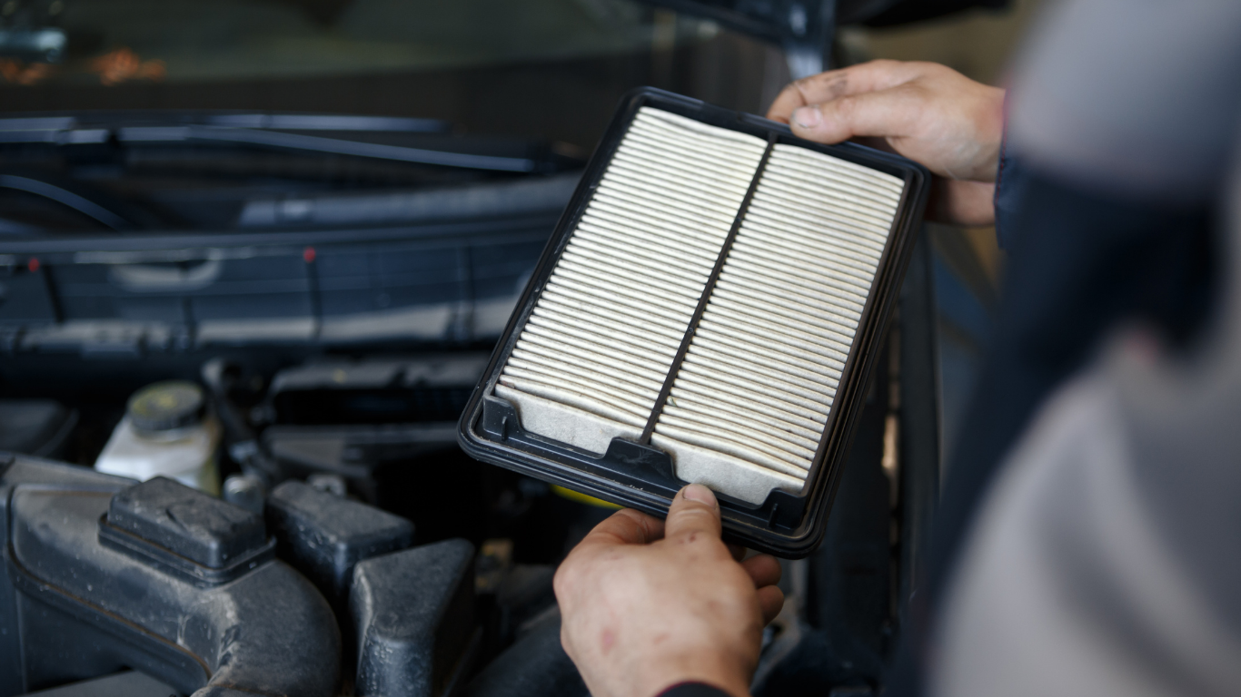 Hands holding a used, rectangular car air filter near an open car hood.