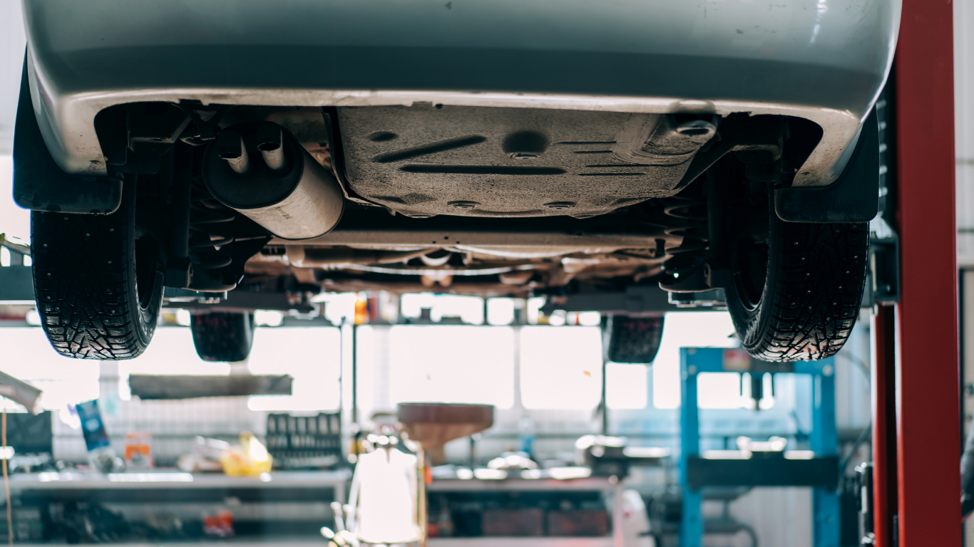 Car raised on a lift, viewed from below in a garage, showing tires, exhaust, and undercarriage.
