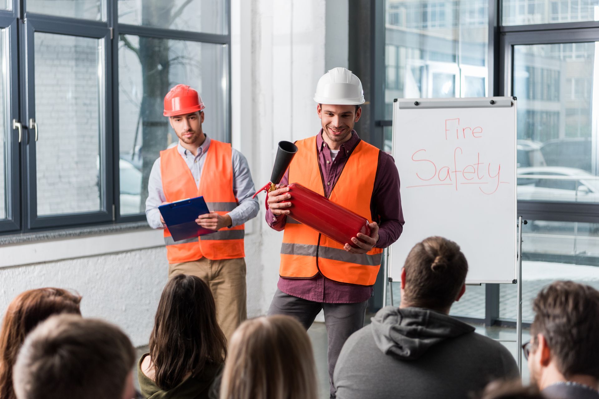 Two men demonstrate fire extinguisher use in a safety training session. Audience seated, whiteboard with