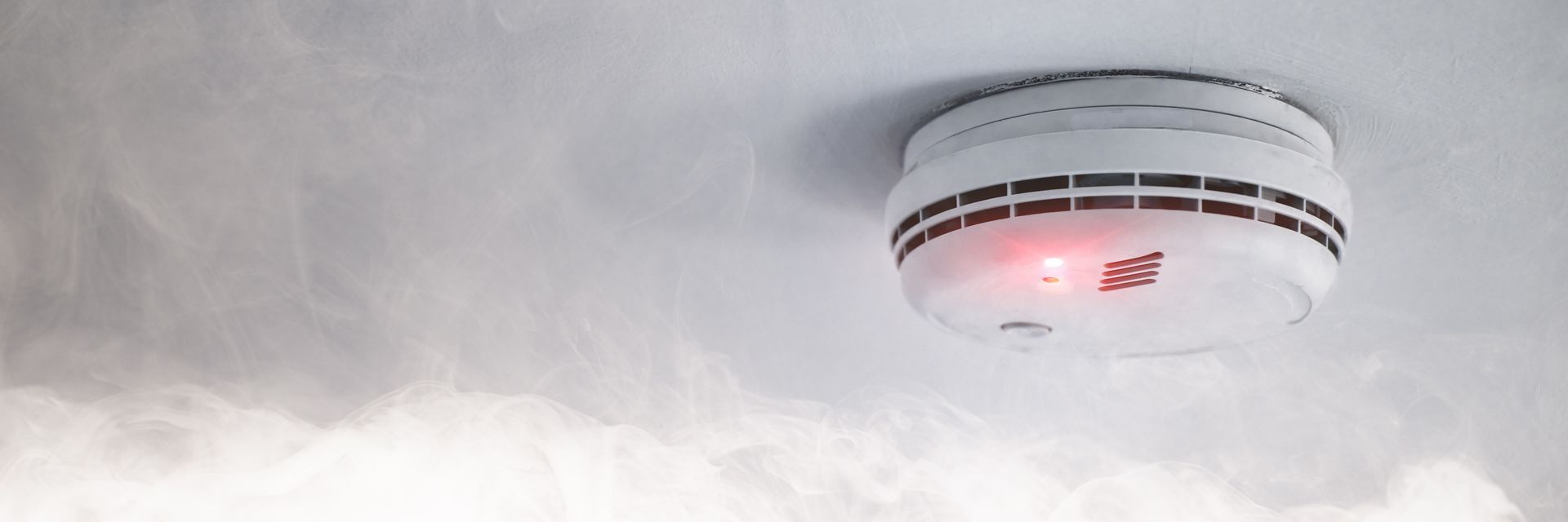 Smoke detector on a ceiling, with a red light indicating activation, and smoke in the room.