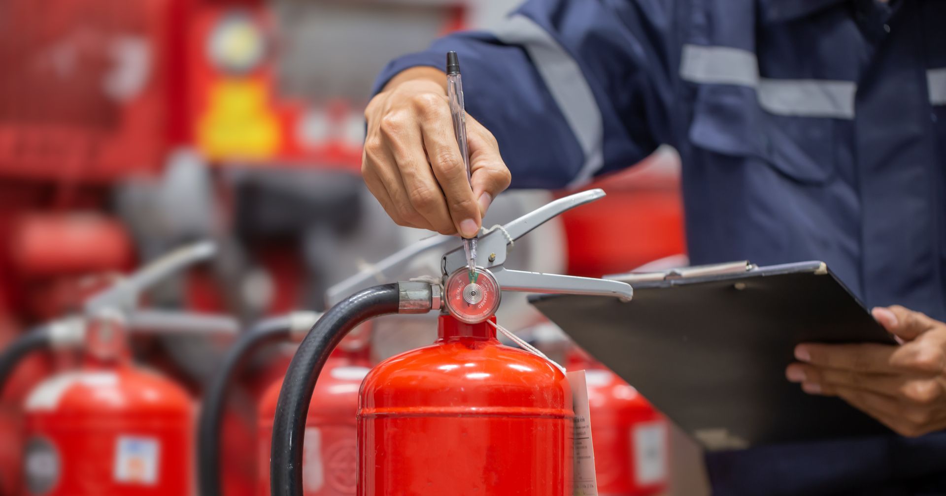 Person inspecting a red fire extinguisher, holding a clipboard, in a room with other extinguishers.