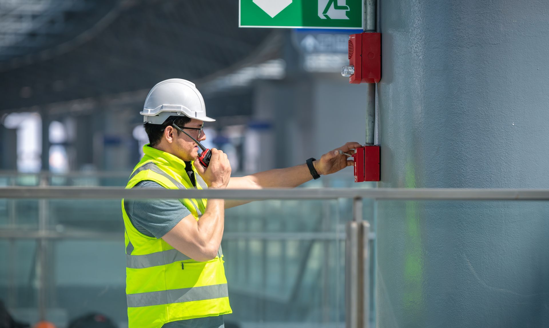 Construction worker in a safety vest and hard hat activating a fire alarm near an exit sign.