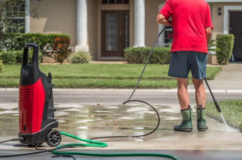 A Man Is Using A High Pressure Washer To Clean A Driveway In Front Of A House — Sadie's Cleaning Services in Koongal, QLD
