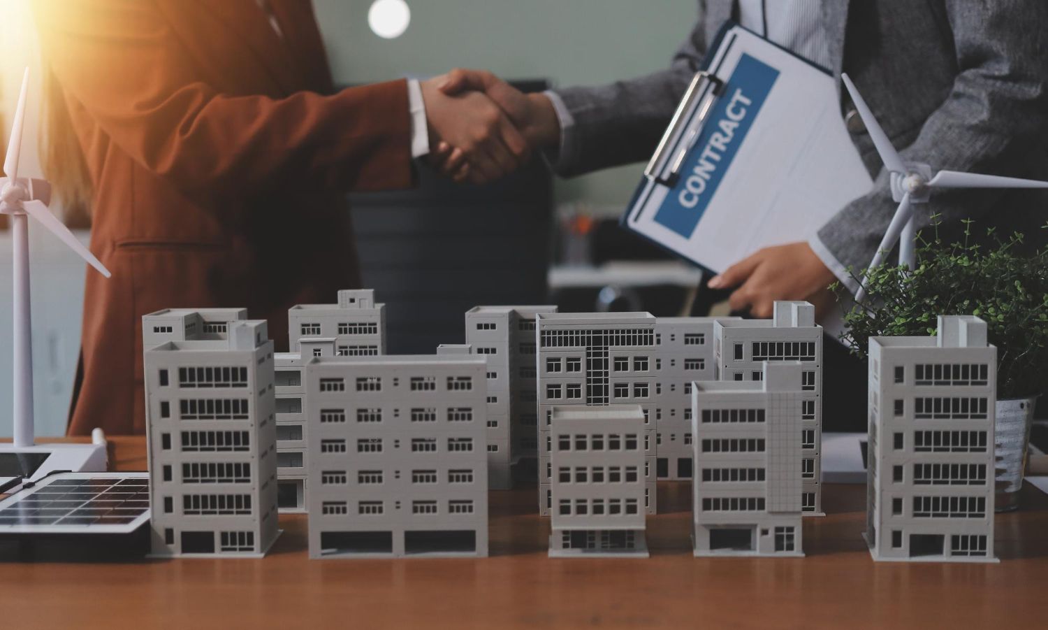 Two people shake hands over model office buildings, with a blue property folder in the background.