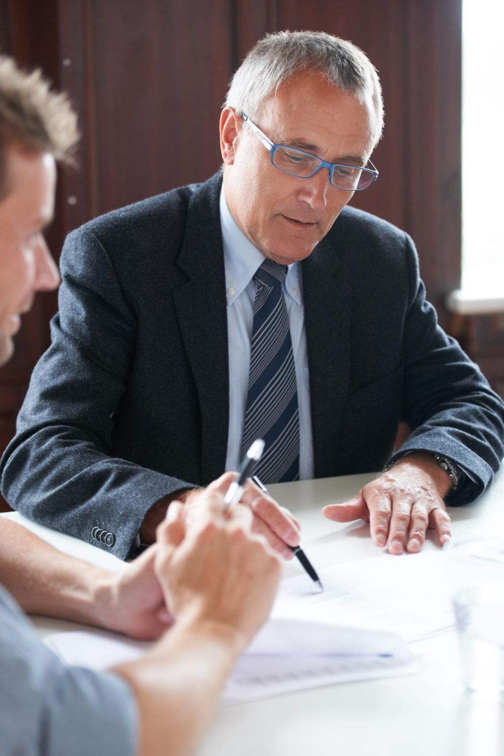 Two people in a meeting; an older man in a suit writes on paper at a conference table.