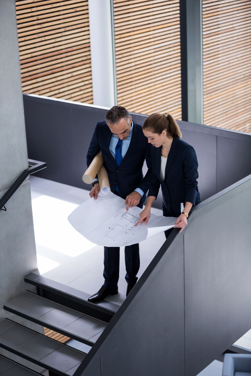 Two coworkers reviewing papers at the top of a stairwell with white walls and wood blinds