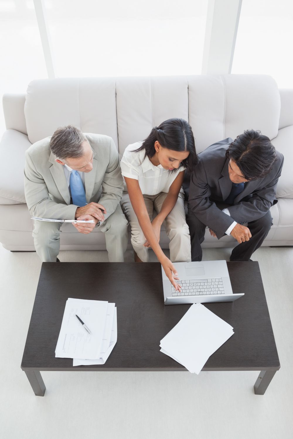 Three coworkers review a laptop at a coffee table in a bright office lounge.