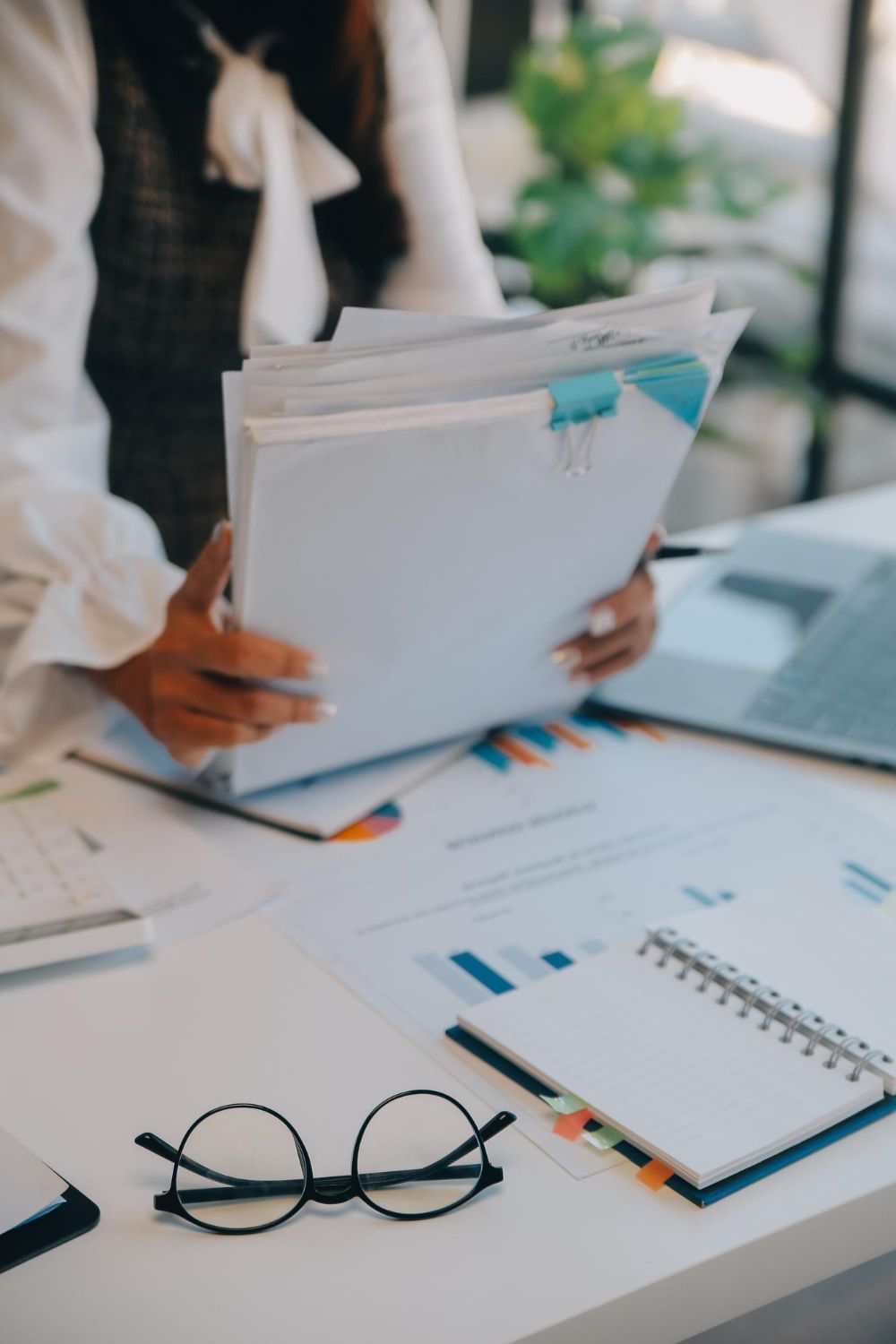 Person reviewing documents at a desk with charts, notebook, glasses, and laptop in an office workspace