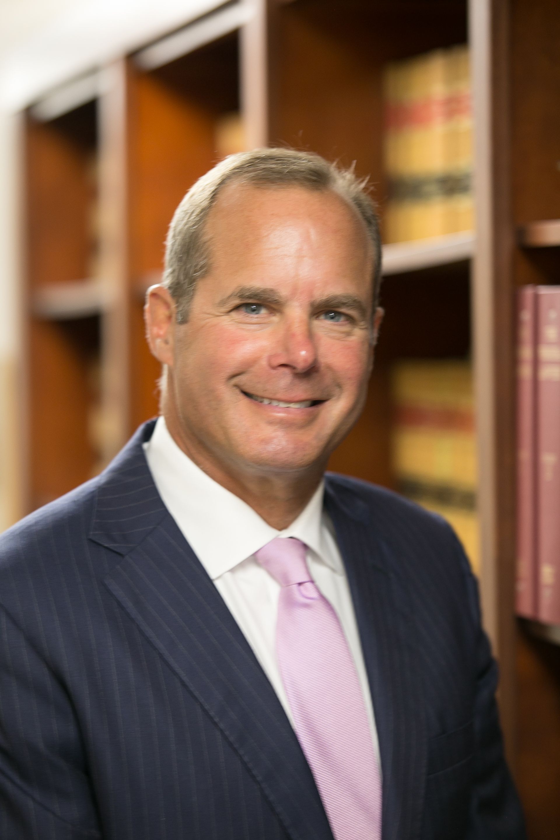 Man in navy suit and pink tie smiles in a library, bookshelves in the background.