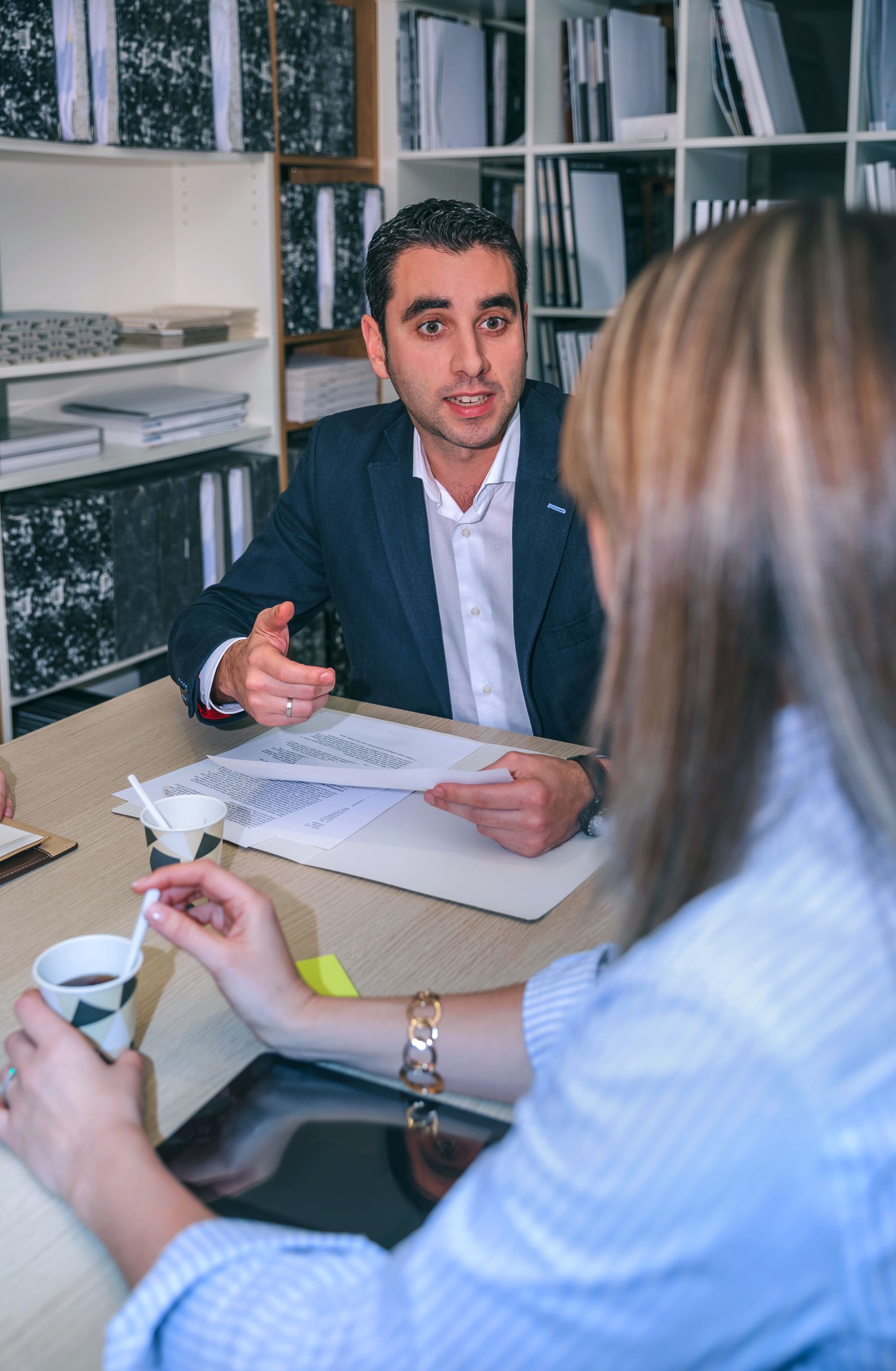 Two people in a meeting, one speaking across a desk with papers and a coffee cup.