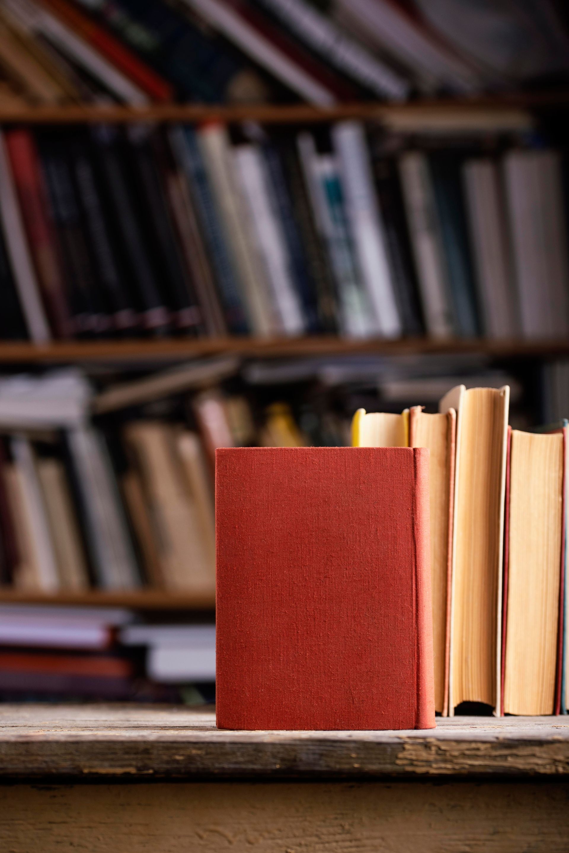 Stack of books on a wooden surface with a red book in front, against a blurred bookshelf background