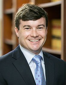 Man with glasses and beard in a business suit smiling at the camera, standing indoors.
