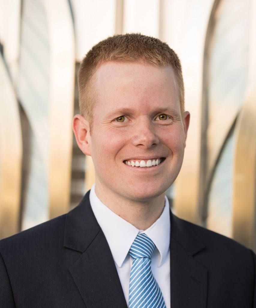Woman smiling, wearing a black blazer and patterned top, in a bright office setting.