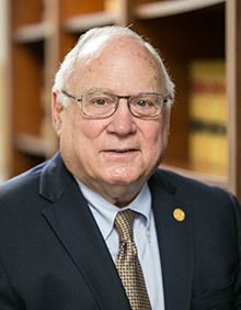 A man with a beard wearing glasses and a suit sits at a desk, looking thoughtful. He is in an office setting.