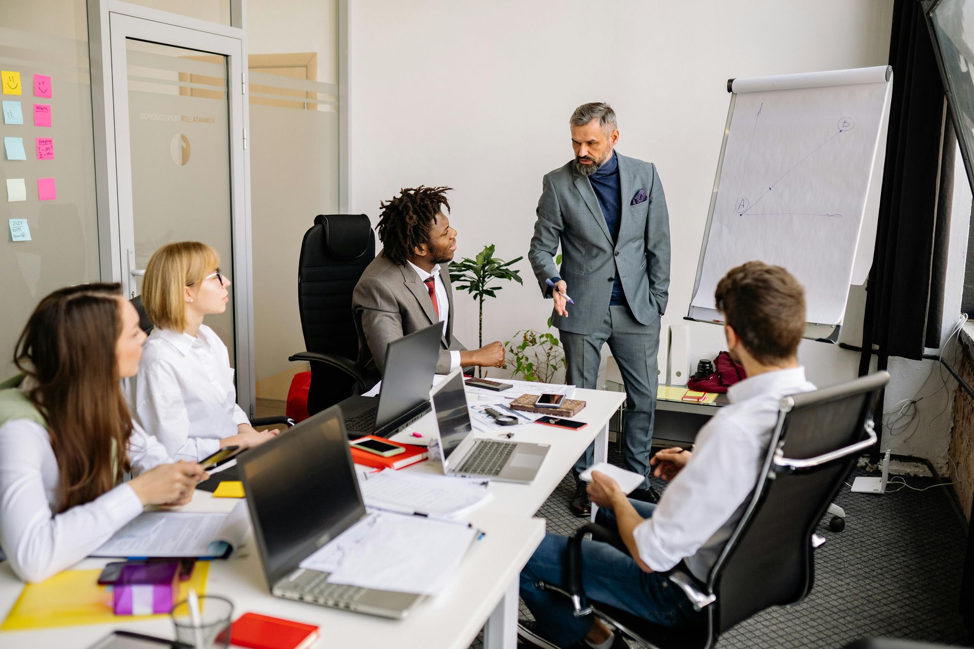 Business meeting in a bright office, with people seated around a table and a presenter beside a flip chart.