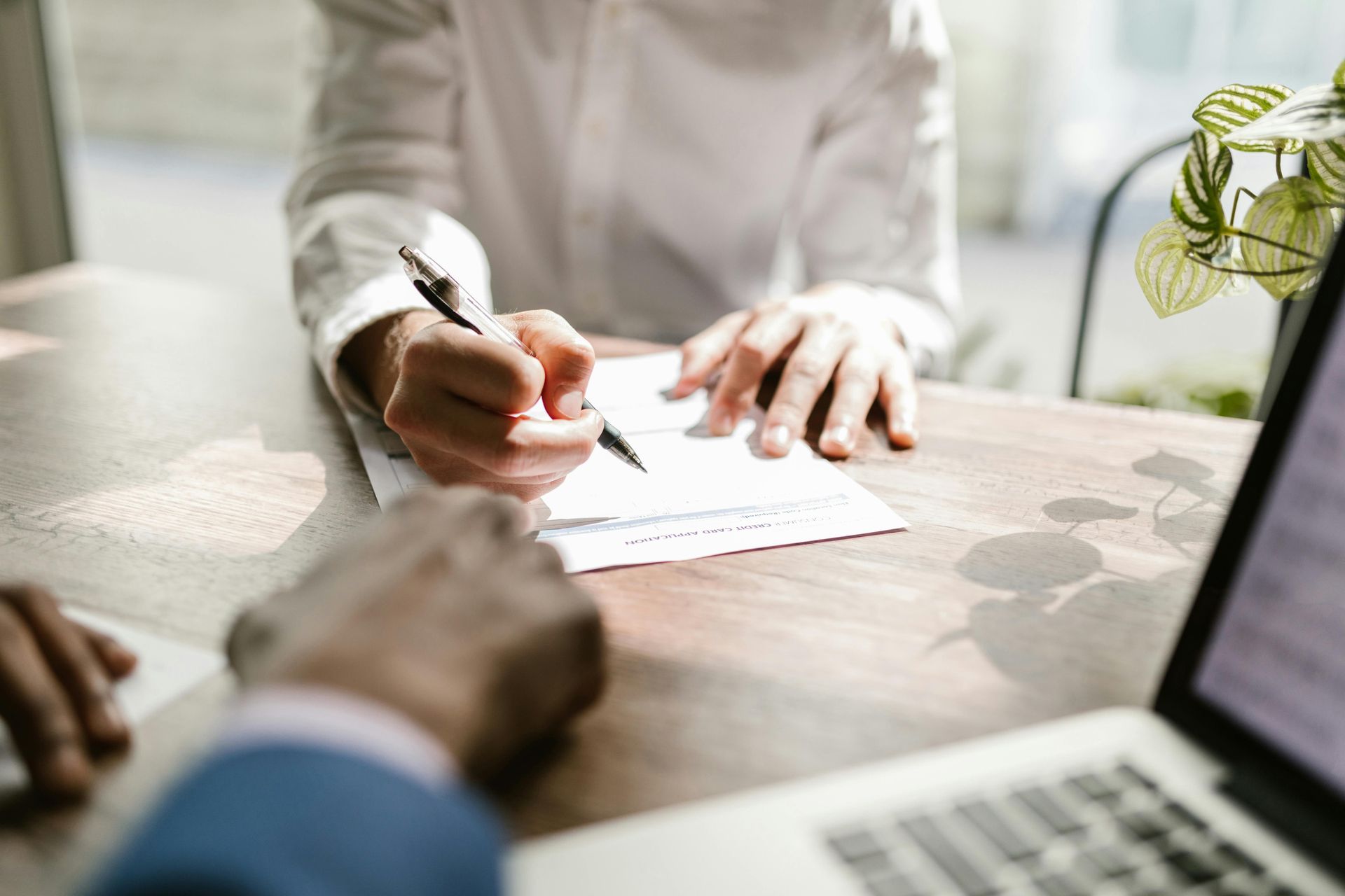 Person signing a document at a table while another points, with a laptop nearby