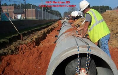 Construction worker in a hard hat and safety vest lowers a large concrete pipe into a trench at a site in NAS Meridian, MS.