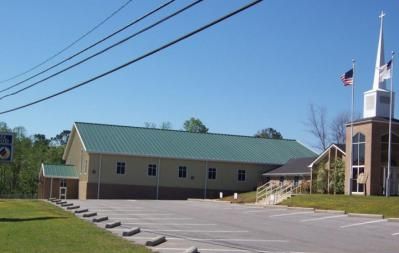 Church building with a green roof and steeple, American flags, and parking lot on a sunny day.