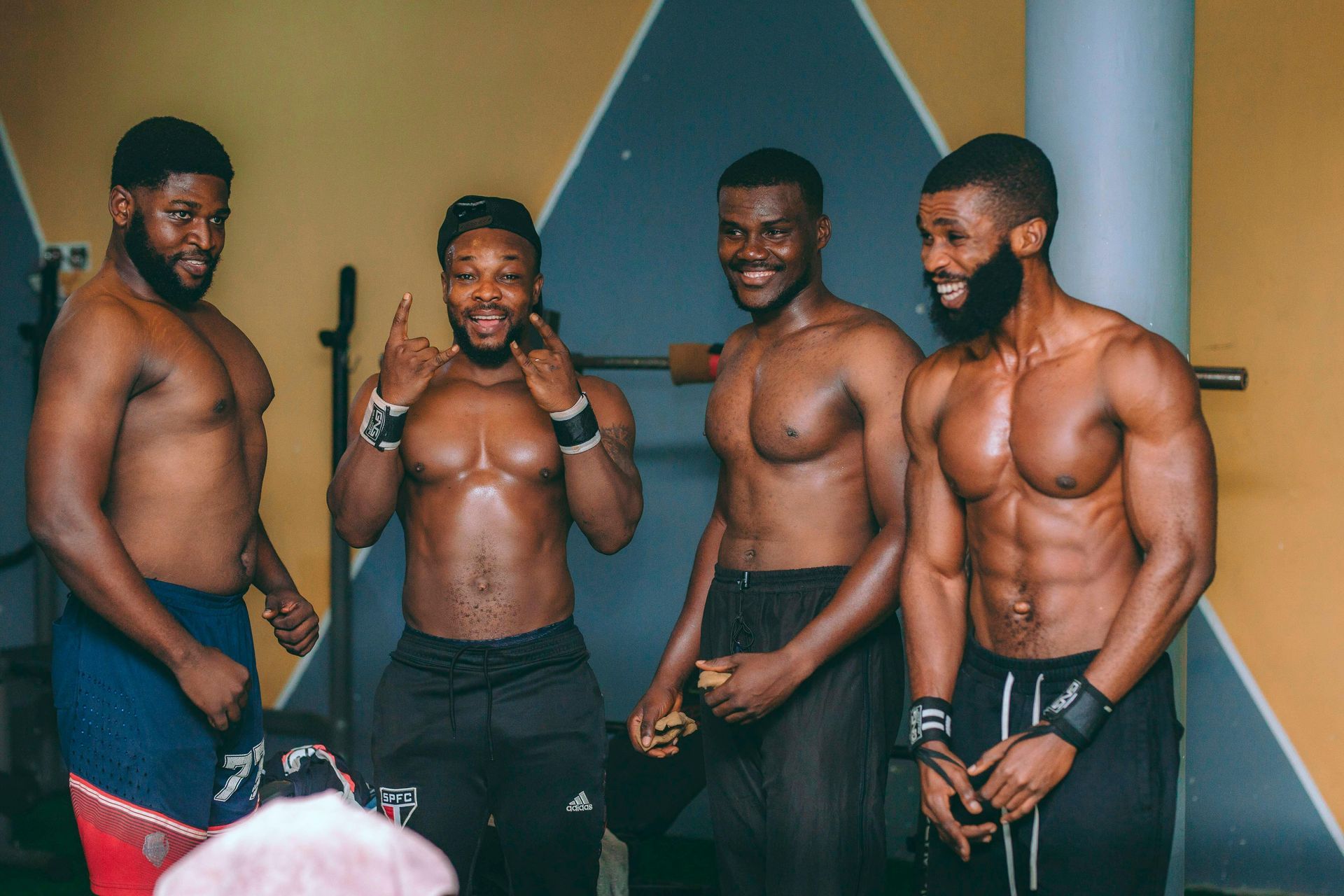 Four shirtless muscular Black men in a gym, smiling, making gestures.