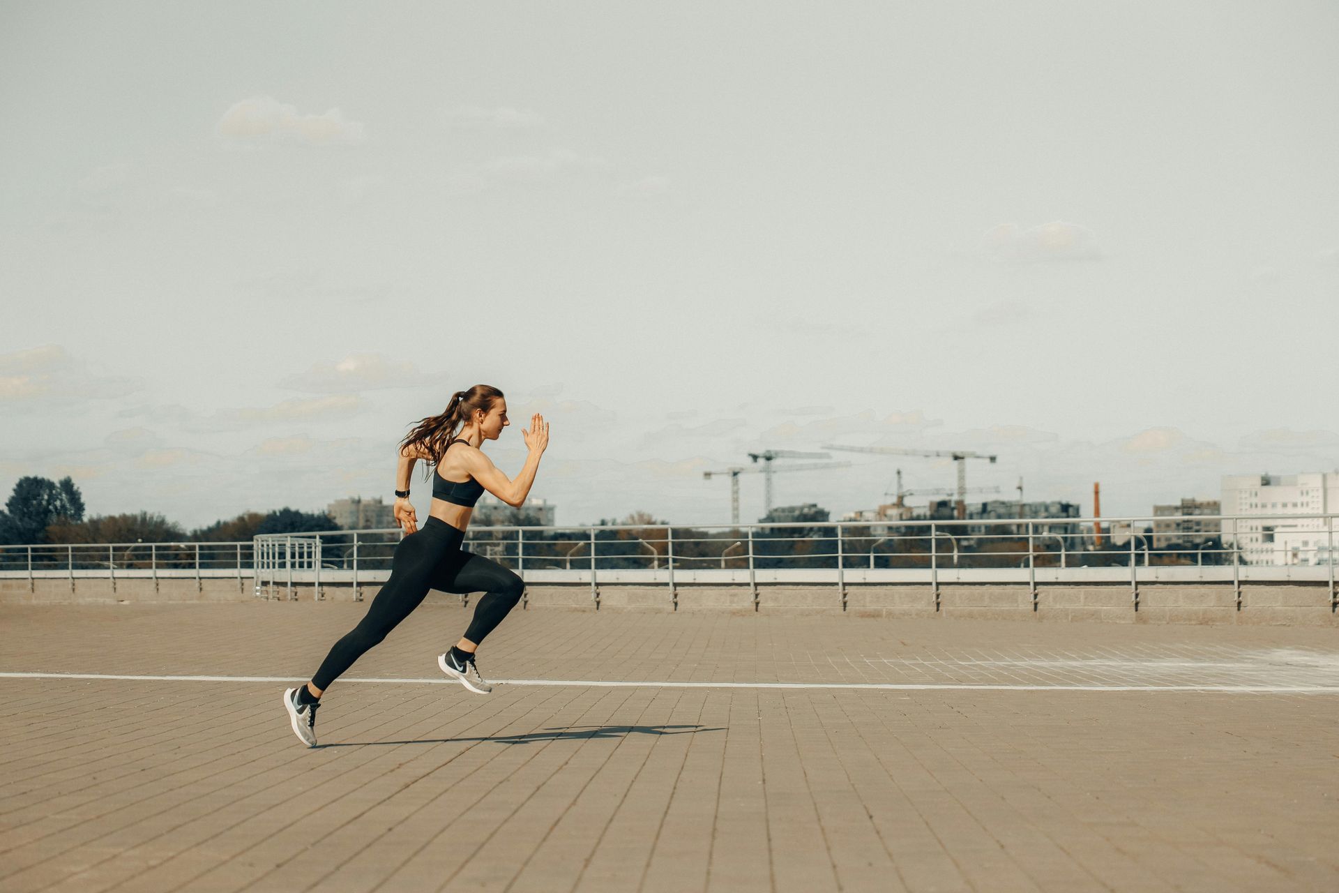 Woman running on a track, wearing black athletic wear. Sky overhead, buildings in the background.