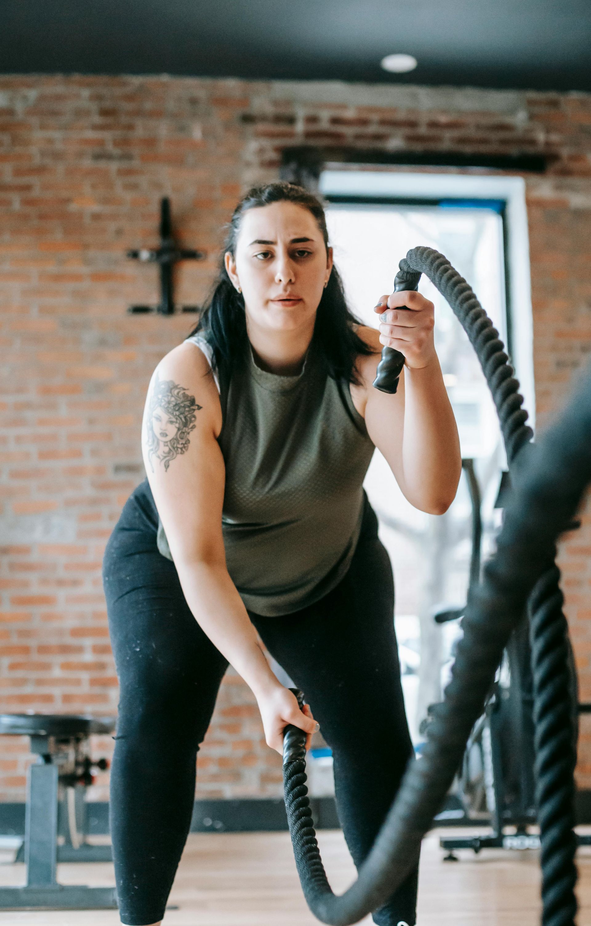 Woman in a gym using battle ropes, looking focused. Brick wall background, dark leggings, green top.