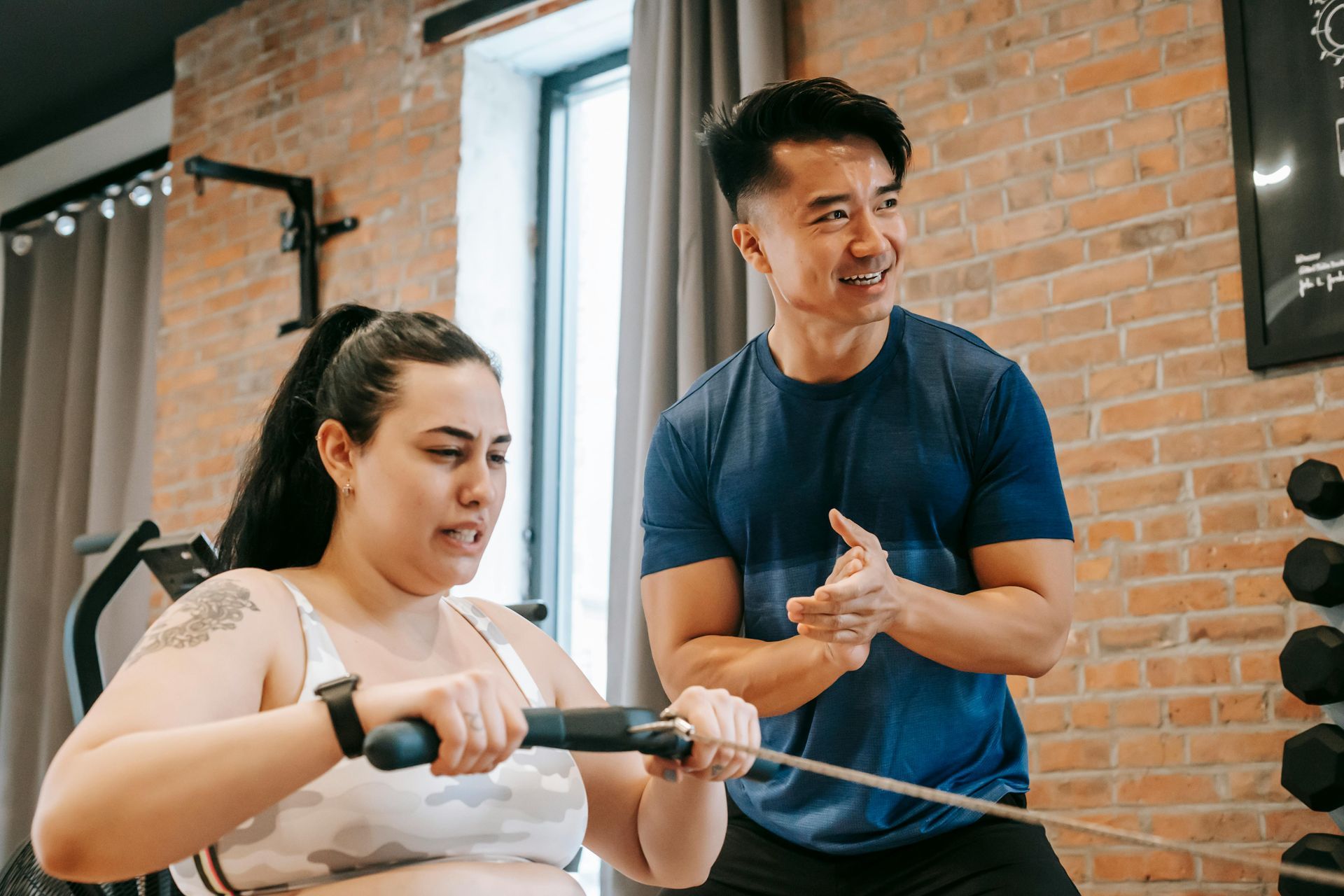 A personal trainer guides a woman using a rowing machine in a brick-walled gym.