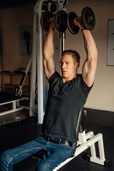 Man lifting dumbbells overhead while seated on weight bench in gym.